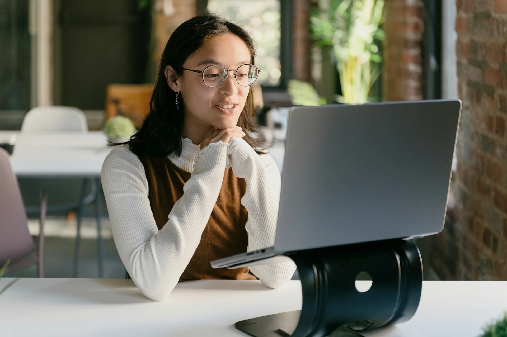A woman with glasses sitting at a desk, smiling while looking at her laptop screen during a video call.